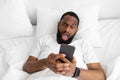 Surprised cheerful millennial african american man with beard and open mouth lies on white bed