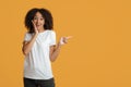 Surprised cheerful young african american female in white t-shirt presses hand to cheek and points to empty space