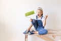 A surprised child Builder sits on a construction ladder in an apartment with white walls and a roller in his hands and shows a Royalty Free Stock Photo