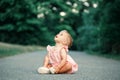 Surprised curious Caucasian happy baby girl sitting on ground empty road outdoor looking up. Funny cute adorable kid child toddler
