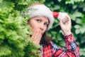 Surprised face woman in a santa claus red hat looks away through green fir tree branches in the forest. Christmas coming, sale