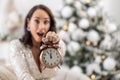 Surprised facial expression of a woman next to a Christmas tree showing alarm clock with five minutes to twelve Royalty Free Stock Photo
