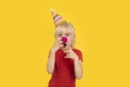 Surprised fair-haired boy with clown nose and party cap on yellow background. Kids party