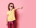 Surprised frolic 6-7 y.o. kid girl in yellow t-shirt, shiny pink leggings and sunglasses pointing finger at copy space