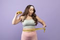 Surprised funny hungry caucasian plus size young lady in sports uniform with burger measures waist with measuring tape