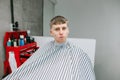 Surprised funny young man sitting in the armchair of a barber shop, wearing a peignoir and looking at the camera with a funny face Royalty Free Stock Photo