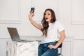 Surprised girl freelancer with a laptop and phone in her hand makes selfie. Young woman with a white T-shirt and jeans Royalty Free Stock Photo