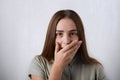 Surprised girl with long straight hair and dark eyes covering her mouth with hand over grey background and looking at camera. A su Royalty Free Stock Photo