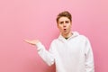 Surprised guy in white hoodie stands on pink background, holds copy space in his hand and looks into the camera. Shocked young man Royalty Free Stock Photo