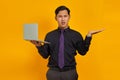 Surprised handsome young businessman showing copy space in palm and holding laptop on yellow background Royalty Free Stock Photo