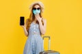 Surprised woman in summer hat and medical mask showing blank smartphone screen while standing with suitcase on yellow background Royalty Free Stock Photo