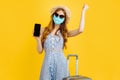 Surprised woman in summer hat and medical mask showing blank smartphone screen while standing with suitcase on yellow background Royalty Free Stock Photo