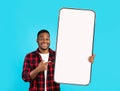 Surprised happy young african american guy in casual points finger at huge smartphone with blank screen