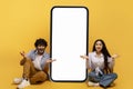 Surprised indian couple sitting near huge smartphone with blank screen over yellow studio background, mockup