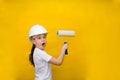 Surprised little girl in a construction white helmet holds a paint roller, standing back on a yellow background