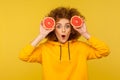 Surprised lively energetic woman with fluffy curly hairstyle holding half slice grapefruit near face, looking at camera