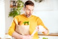 Surprised man looking at store receipt after shopping, holding a paper bag with healthy food. Guy in the kitchen