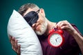 Surprised man in red t-shirt and sleep mask on head holding alarm clock and pillow. Lifestyle and bed time concept Royalty Free Stock Photo
