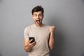 Surprised man with short dark hair shouting and holding smartphone with clenching fist, isolated over gray background Royalty Free Stock Photo