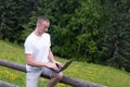 Surprised man sits on a wooden fence and works behind a laptop near the field and coniferous forest Royalty Free Stock Photo