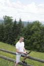 Surprised man sits on a wooden fence and works behind a laptop near the field and coniferous forest. Vertical frame Royalty Free Stock Photo