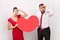 Surprised man and woman standing together, holding big heart, looking far, waiting holiday. Royalty Free Stock Photo
