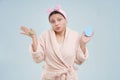 Surprised mid-aged Asian woman wearing a bathrobe, struggling to make choice between skin care products on blue Royalty Free Stock Photo