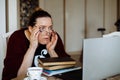 Surprised middle-aged woman sitting at table near books, looking at laptop, adjusting glasses, not believing eyes. Royalty Free Stock Photo