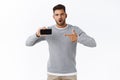 Surprised overwhelmed young handsome man in grey sweater asking question about mobile game or application, holding