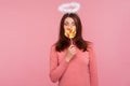 Surprised playful brunette woman with angelic halo holding big colorful lollipop near mouth, having fun eating rainbow candy