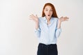 Surprised redhead woman looking down with hands spread sideways in disbelief, standing shocked against white background Royalty Free Stock Photo