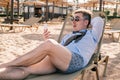 Surprised relaxed businessman downshifter lies on a sun lounger on the beach looking at the phone, wears shirt, tie and