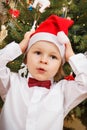 Surprised or shocked baby boy dressed in festive clothing and Santa Claus hat. Christmas tree with decoration in background Royalty Free Stock Photo