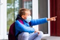 Surprised side view wearing face mask young schoolboy sitting on a stairs near school door and pointing finger somewhere Royalty Free Stock Photo