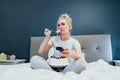 Surprised, stressed, shocked woman eating cake and holding a remote control while watching TV in bed. Scary horror movie Royalty Free Stock Photo