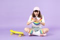 Surprised teen girl in vivid clothes sitting near skateboard using mobile phone typing sms message isolated on violet Royalty Free Stock Photo