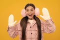 surprised teen girl in winter mittens and earmuffs on yellow background, winter fashion Royalty Free Stock Photo