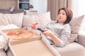 Surprised woman eating pizza and watching TV with remote control at home, warm tone Royalty Free Stock Photo
