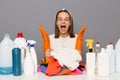 Surprised woman in orange rubber gloves posing at workplace with cleaning detergents isolated over gray background screaming with