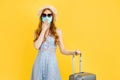 Surprised woman traveler, wearing a medical mask, wearing a hat, with a suitcase on a yellow background Royalty Free Stock Photo