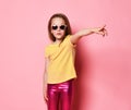 Surprised 6-7 y.o. kid girl in yellow t-shirt, shiny pink leggings and sunglasses pointing finger at side