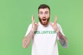 Surprised young bearded man in white volunteer t-shirt isolated on pastel green wall background. Voluntary free work