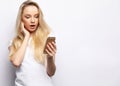 Surprised young blond hair woman holding mobile phone and staring at it while standing against white background. Royalty Free Stock Photo