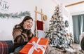 Surprised young caucasian woman with big Christmas present while sitting on a bed at home near Christmas tree Royalty Free Stock Photo