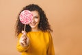 Surprised curly girl eating lollipop. Beauty Model woman holding pink sweet colorful lollipop candy, isolated on beige background Royalty Free Stock Photo