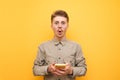Surprised young man in glasses and shirt and mustache stands on yellow background with smartphone in hand, looks into camera with Royalty Free Stock Photo