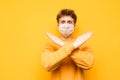 Surprised young man in a protective mask stands on a yellow background and shows his hands the cross. The guy in a medical mask