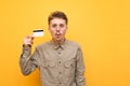 Surprised young man in shirt and glasses stands on yellow background with bank card in hand, looks into camera with surprised face Royalty Free Stock Photo