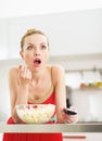 Surprised young woman eating popcorn and watching tv in kitchen Royalty Free Stock Photo
