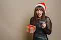 Surprised young woman in santa claus hat on a beige background holding a Christmas present with a red ribbon and a credit card Royalty Free Stock Photo